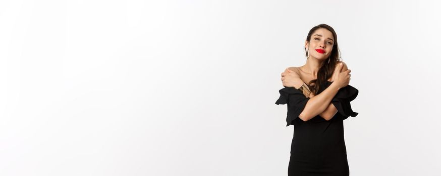 Fashion and beauty. Sensual and tender woman in black dress, embracing own body, hugging herself and smiling, standing over white background.