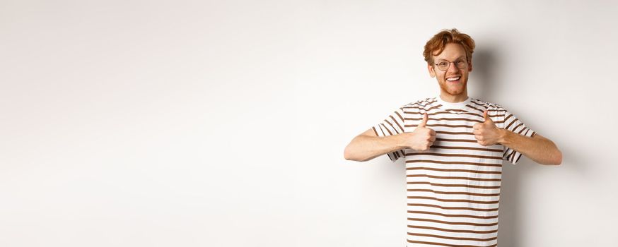 Cheerful redhead guy in nerdy glasses showing thumbs-up, smiling and saying yes, agree or like something, standing over white background.