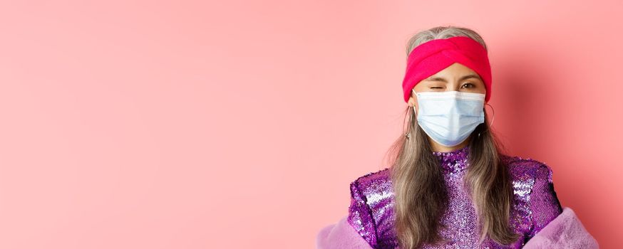 Covid-19, virus and social distancing concept. Sassy asian senior woman in medical mask and glitter dress winking, wearing party outfit, pink background.