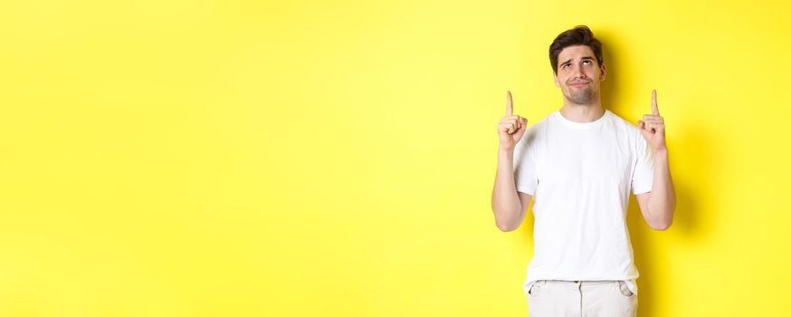 Skeptical young man pointing and looking up at something bad, judging offer, standing over yellow background.