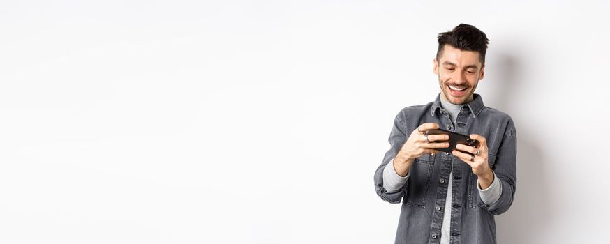 Handsome young man playing mobile video game and smiling at screen, standing against white background.