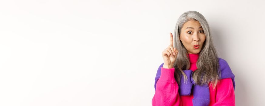 Close up of elderly asian woman in stylish hipster clothes, raising finger and saying idea, have suggestion, standing over white background.