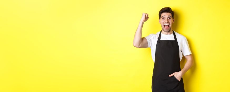 Cheerful seller making fist pump, rejoicing and triumphing, standing in black apron against yellow background.