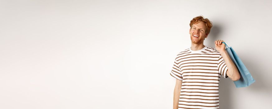 Cheerful bearded man with red hair enjoying shopping, holding bag over shoulder and smiling, standing over white background.