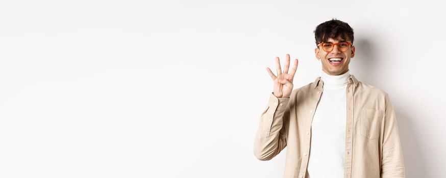 Handsome young man making order, showing number four fingers and smiling, standing on white background.