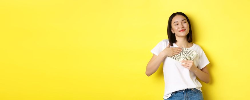 Beautiful asian woman love money, hugging dollars and smiling pleased, standing over yellow background.