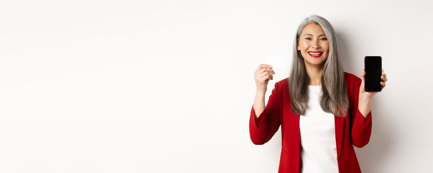 Senior asian businesswoman showing plastic credit card and blank smartphone screen, smiling at camera, white background.