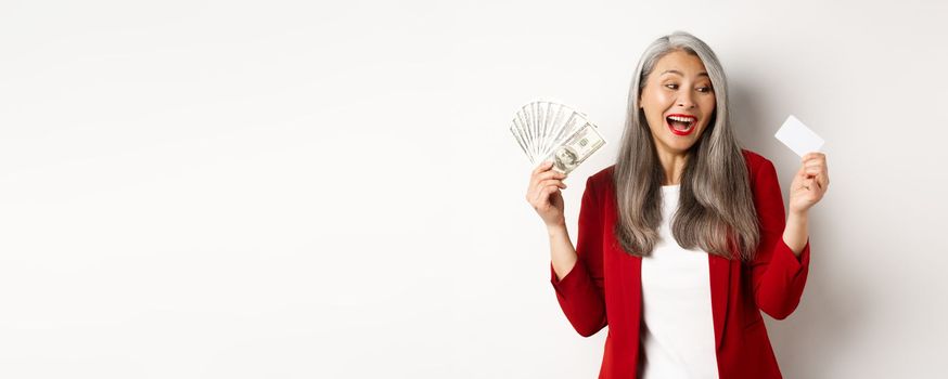 Cheerful asian senior woman earn money, showing dollars in cash and looking at plastic credit card, smiling excited, standing over white background.