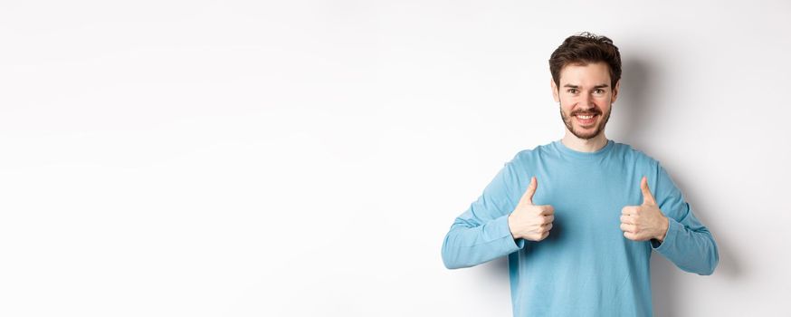 Smiling handsome man showing thumbs up, like and approve good choice, praising or recommending something, standing on white background.