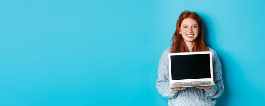 Young smiling woman with red hair and freckles showing computer screen, holding laptop and demonstrate online promo, standing over blue background.