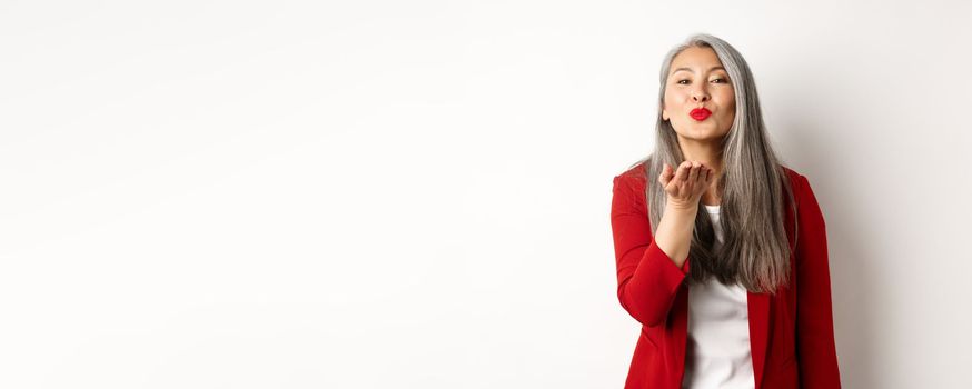 Elegant asian woman with red blazer and lips, blowing air kiss at camera, concept of valentines day and romance, white background.