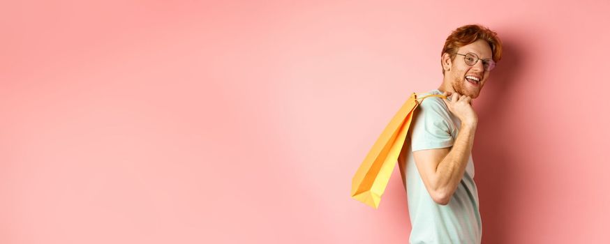 Carefree young man with red hair and glasses, walking with shopping bag over his shoulder and smiling, shopper buying presents, standing satisfied over pink background.