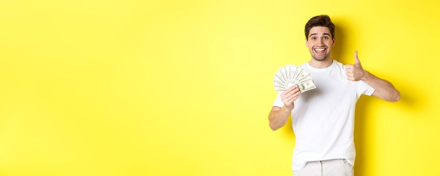 Satisfied buyer man showing thumb-up and holding money, shopping with cash, standing over yellow background.