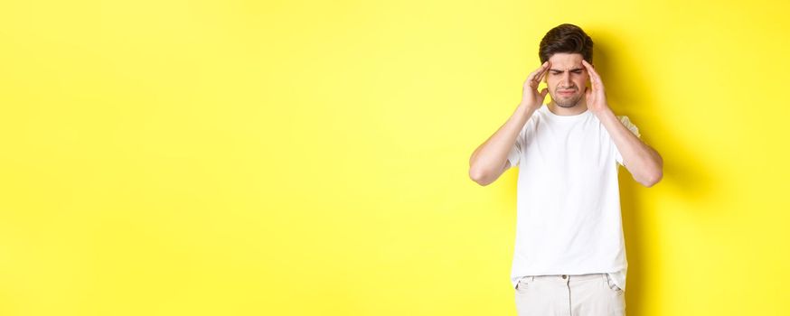 Troubled man touching head and grimacing from pain, complaining on headache, standing over yellow background. Copy space