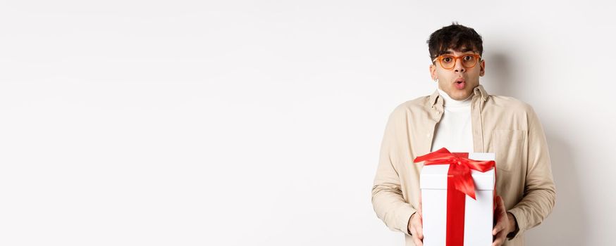 Surprised young man say wow and holding gift box, receive present on holiday, standing amused on white background.