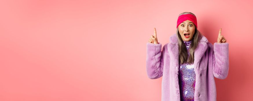 Happy elderly asian lady pointing fingers up, looking amazed at camera, showing cool promo offer, standing against pink background in purple fake fur coat.