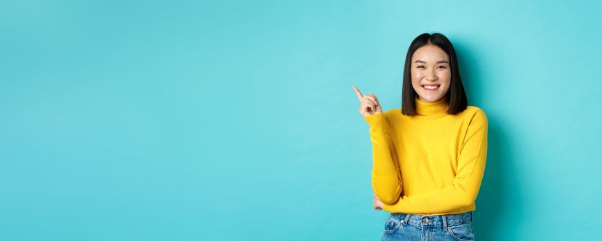 Image of beautiful young asian woman with perfect smile and no blemishes on skin, pointing finger left, looking satisfied, standing over blue background.