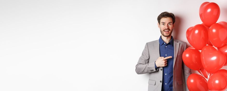 Lovers day. Excited male model in suit pointing finger at Valentines heart balloons and smiling, prepare romantic gifts on date, standing over white background.