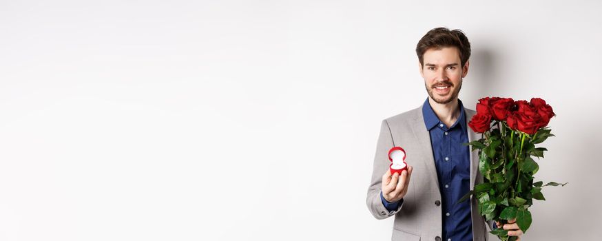 Smiling boyfriend making a marriage proposal, standing with engagement ring and bouquet of red roses, going on romantic Valentines day date, white background.