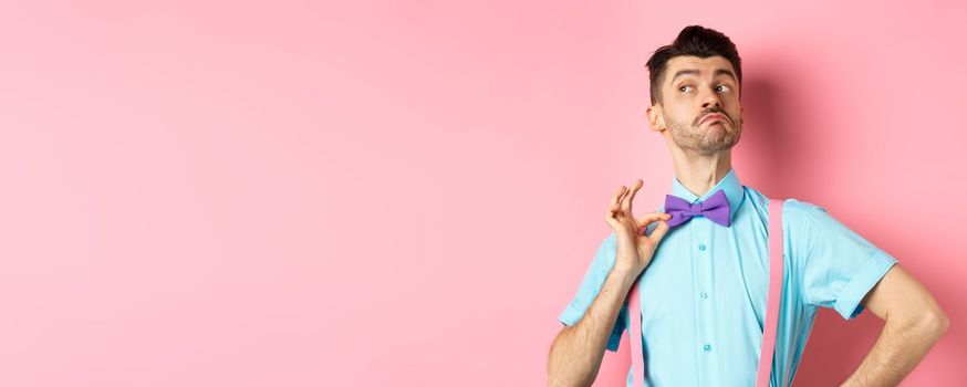 Image of funny man raising chin up and adjusting bow-tie, standing confident and sassy on pink background, looking left.