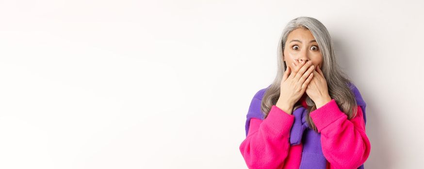 Close up of shocked senior lady gasping and covering mouth, staring at camera speechless, witness something, standing over white background.