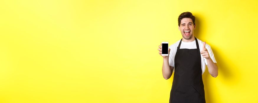Happy waiter showing mobile screen and thumb up, recommending cafe restaurant app, standing over yellow background.
