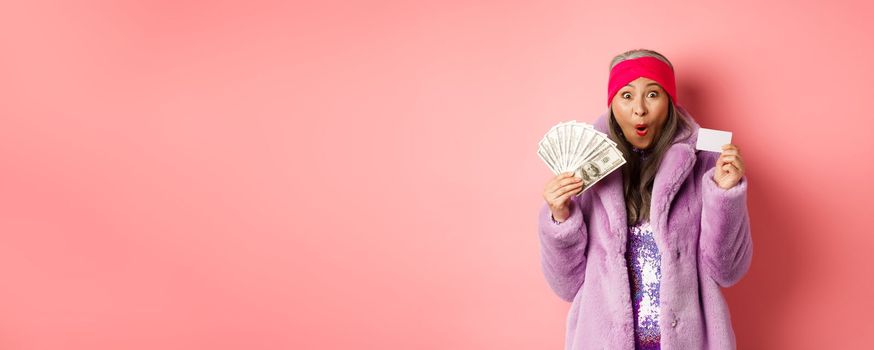 Shopping and fashion concept. Happy senior asian woman holding money in dollars and plastic card, looking amazed and ecstatic, pink background.