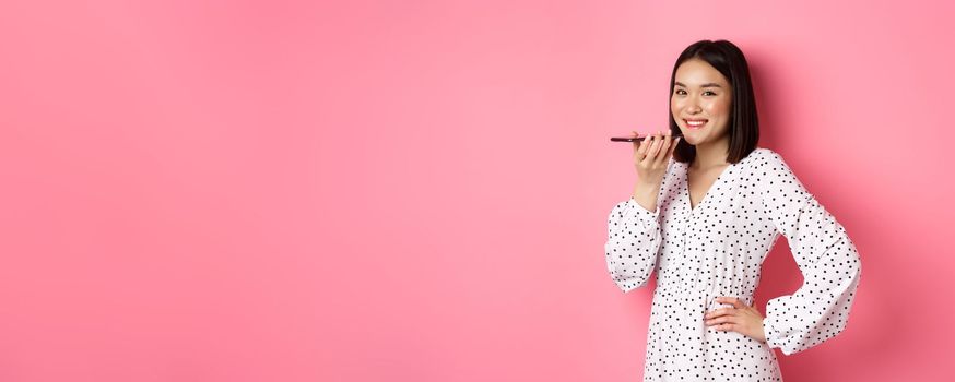 Beautiful korean woman talking on speakerphone, recording voice message and smiling happy, standing over pink background.