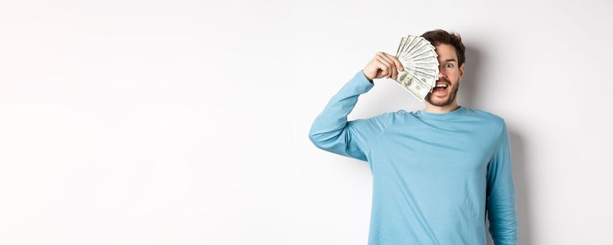 Surprised and happy young man covering half of face with money, gasping amazed at camera, checking out quick loan offers, standing over white background.
