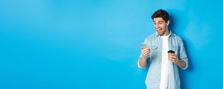 Man looking surprised at smartphone, drinking coffee, standing over blue background amazed.