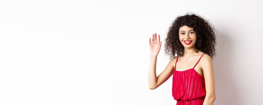 Beauty and fashion. Friendly elegant woman with curly hair, red dress, waving hand and saying hello, smiling to greet you, standing on white background.