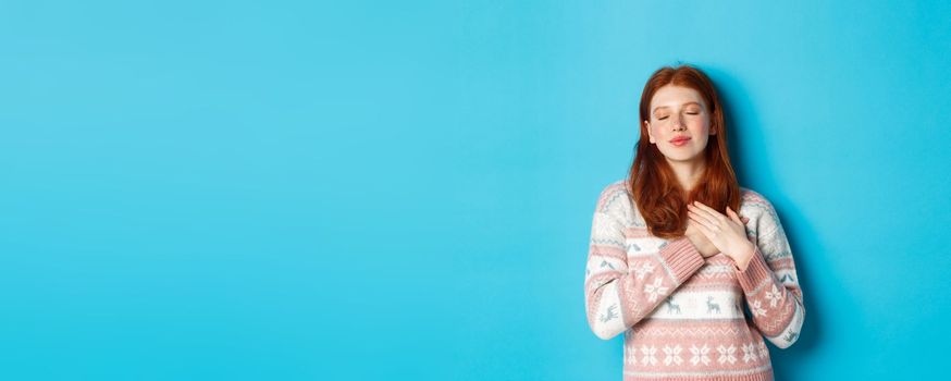 Image of dreamy redhead girl close eyes and holding hands on heart, feeling nostalgic, remember or imagine something, standing over blue background.