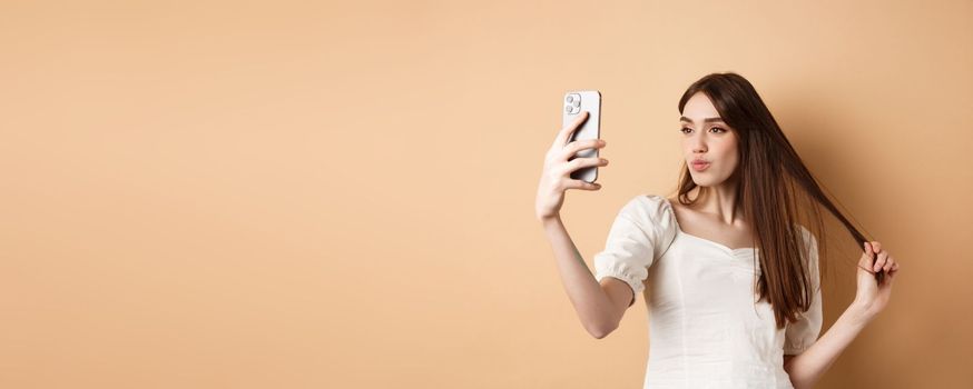 Stylish young woman playing with hair and taking selfie on smartphone, make photo for social media, standing on beige background.