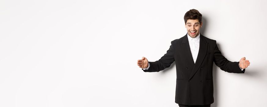 Portrait of excited handsome man in suit, shaping big object on copy space and smiling amazed, holding something, standing over white background.