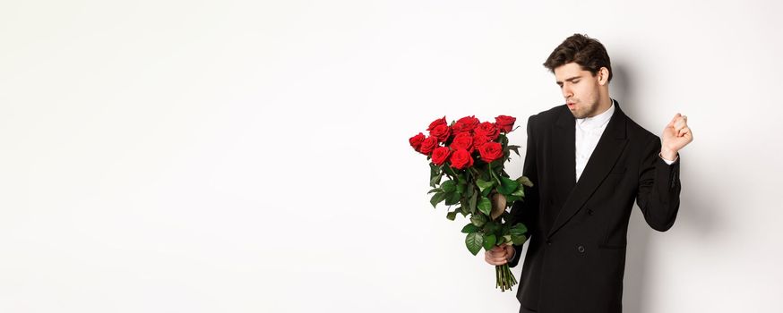 Image of elegant and sassy man in black suit, looking confident and holding bouquet of red roses, going on a romantic date, standing against white background.