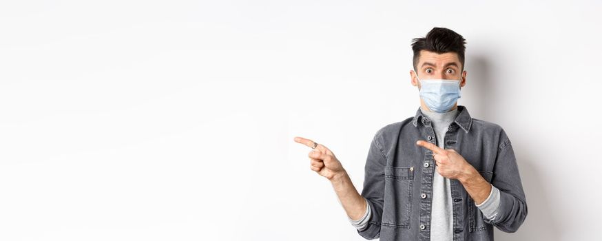 Pandemic lifestyle, healthcare and medicine concept. Young guy in face mask inviting look here, pointing fingers left at logo, standing on white background.