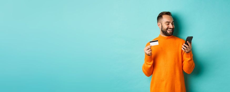 Online shopping. Handsome bearded man paying in internet, holding credit card and stare at mobile screen, standing over turquoise background.