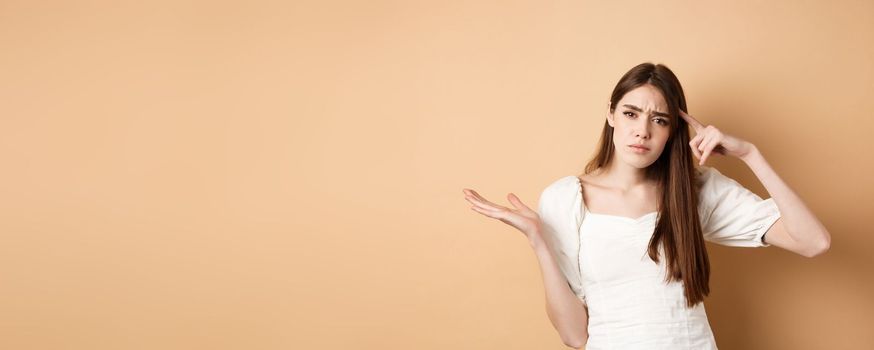 Are you stupid. Annoyed woman mocking person, pointing finger at head and shrugging confused, irritated by crazy ideas, standing on beige background.