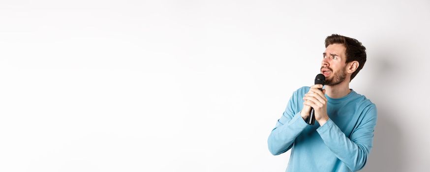 Worried and confused man reading lyrics on karaoke, looking left with unsure face, holding microphone and singing, white background.