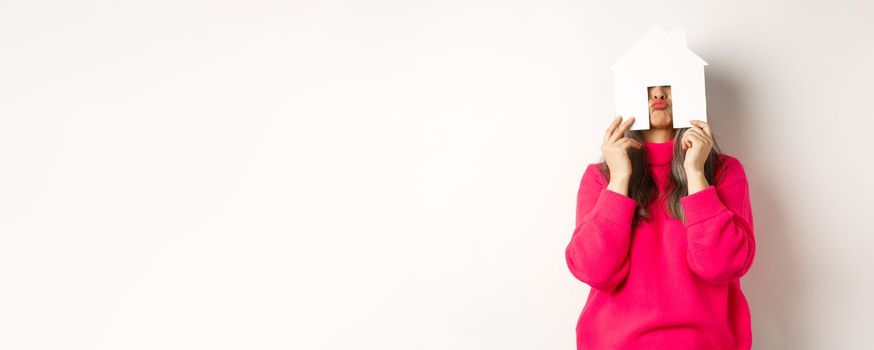 Real estate. Funny asian middle-aged woman hiding face behind paper house model and showing puckered lips, having fun, standing over white background.