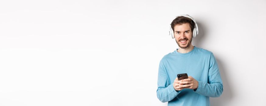 Attractive smiling man listening music in wireless headphones, using black smartphone and looking pleased, white background.