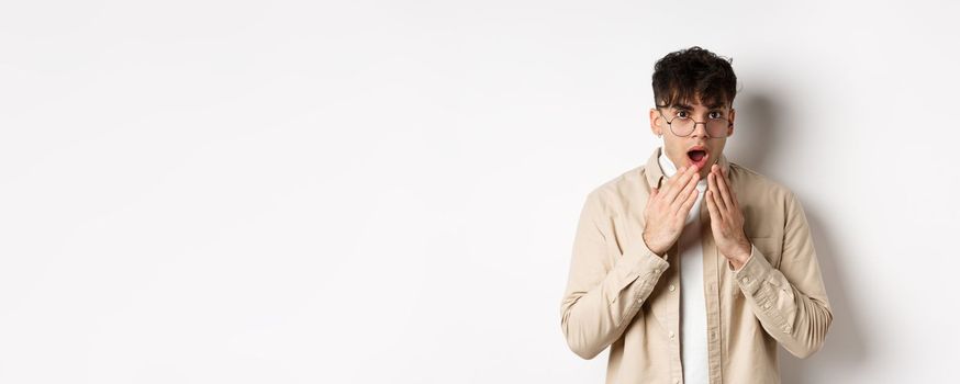 Portrait of handsome guy student looking shocked, hear gossip and stare with disbelief, covering opened mouth with hands, standing on white background.