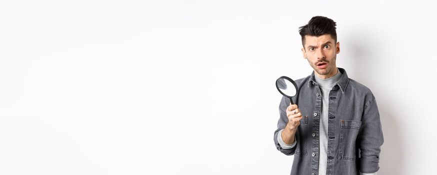Shocked man found something with magnifying glass, look at camera, standing against white background.