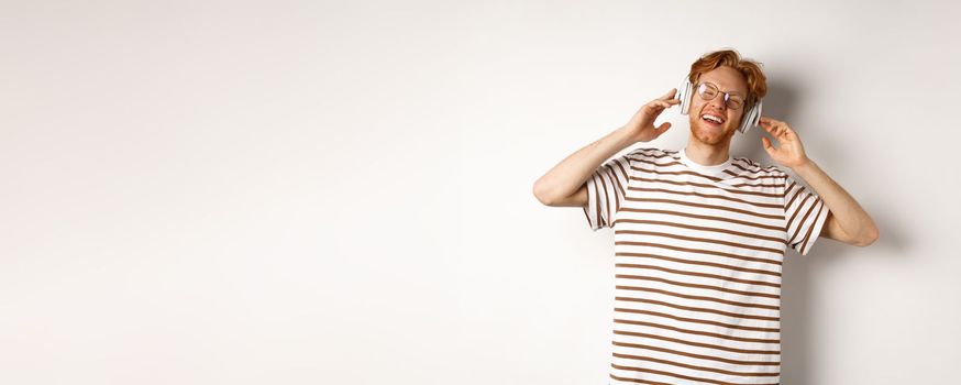 Technology concept. Happy redhead man listening music in headphones and singing along, standing over white background.