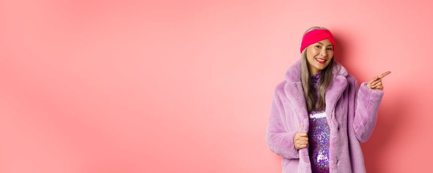 Smiling senior woman looking trendy in winter fur coat, pointing finger right and showing advertisement, standing against pink background.