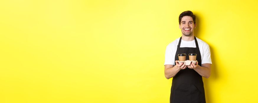 Friendly barista in black apron giving takeaway order, holding two cups of coffee and smiling, standing over yellow background.