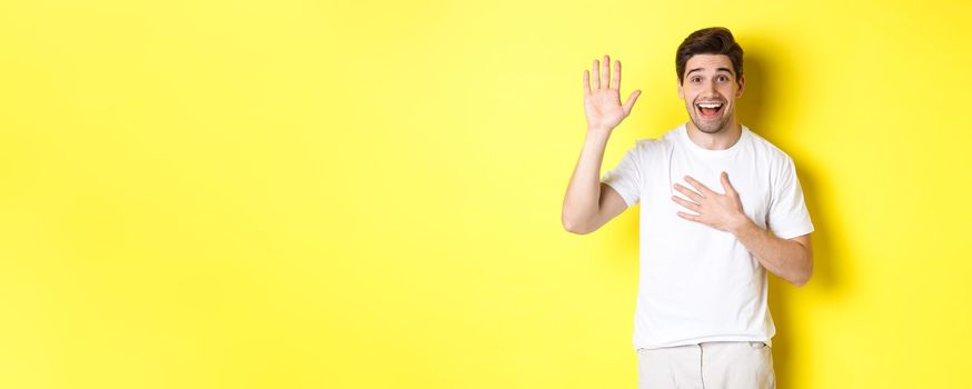 Happy guy making promise, holding hand on heart, swearing to tell truth, standing over yellow background in white t-shirt.