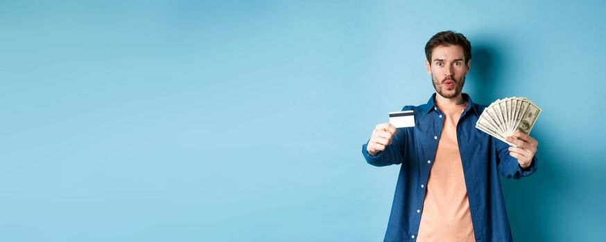 Excited modern guy showing money and plastic credit card, saying wow amazed, standing on blue background.