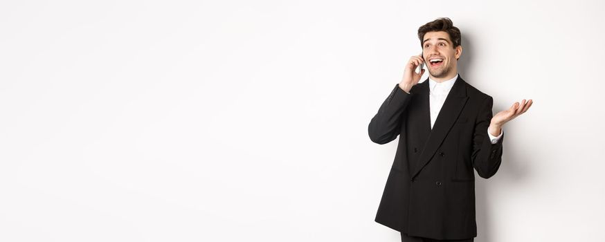 Portrait of happy good-looking businessman receiving great offer, talking on phone and looking pleased, standing in black suit against white background.