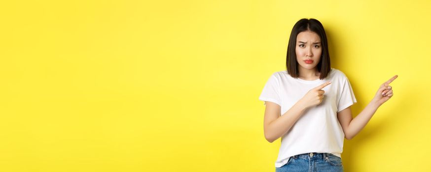 Beauty and fashion concept. Beautiful asian woman in white t-shirt pointing fingers right, demonstrate logo standing over yellow background.